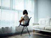 Depression Comedy woman sitting on black chair in front of glass-panel window with white curtains